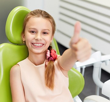 A young girl sitting in a dentist’s chair and pointing her arm out to give a thumbs up