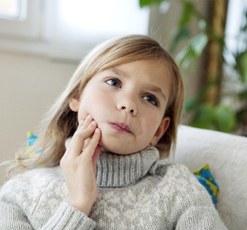 A little girl wearing a sweater and sitting on a couch holding her jaw in pain and needing an emergency dentist in Fitchburg