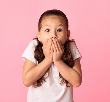 A little girl wearing a t-shirt and holding her hands over her mouth in shock