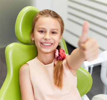A young girl wearing a side ponytail and giving a thumbs up while seated in the dentist’s chair